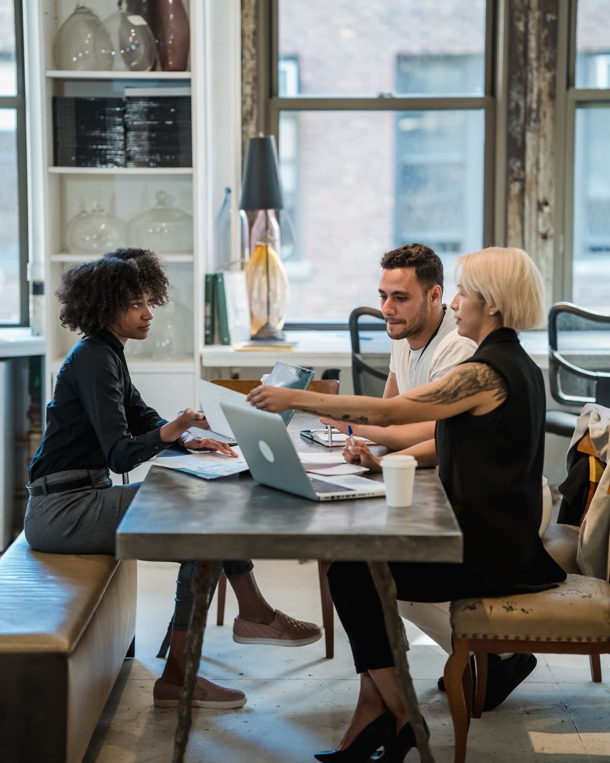 Three people seated at a metal table in a sunlit loft office, talking and passing papers beside a laptop.