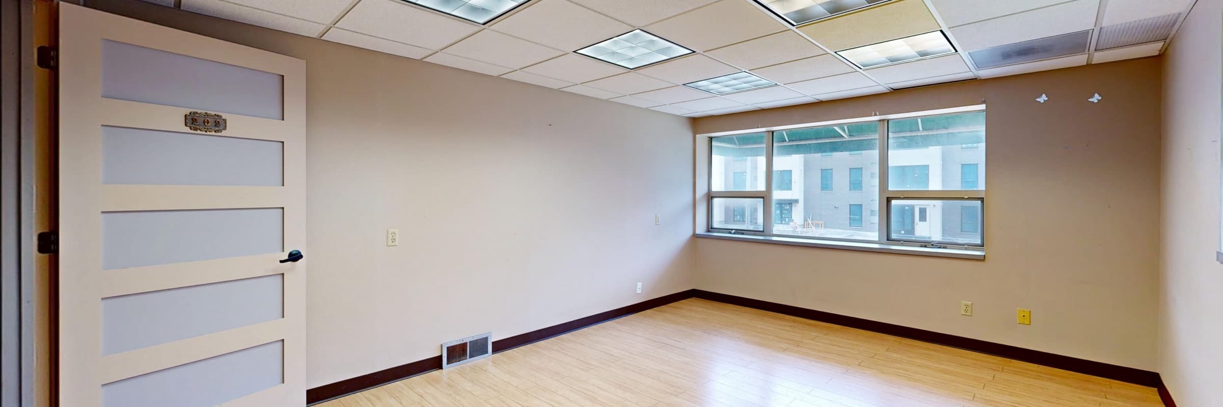 An empty office with a wood floor, a paneled door, ceiling lights, and a wide window.